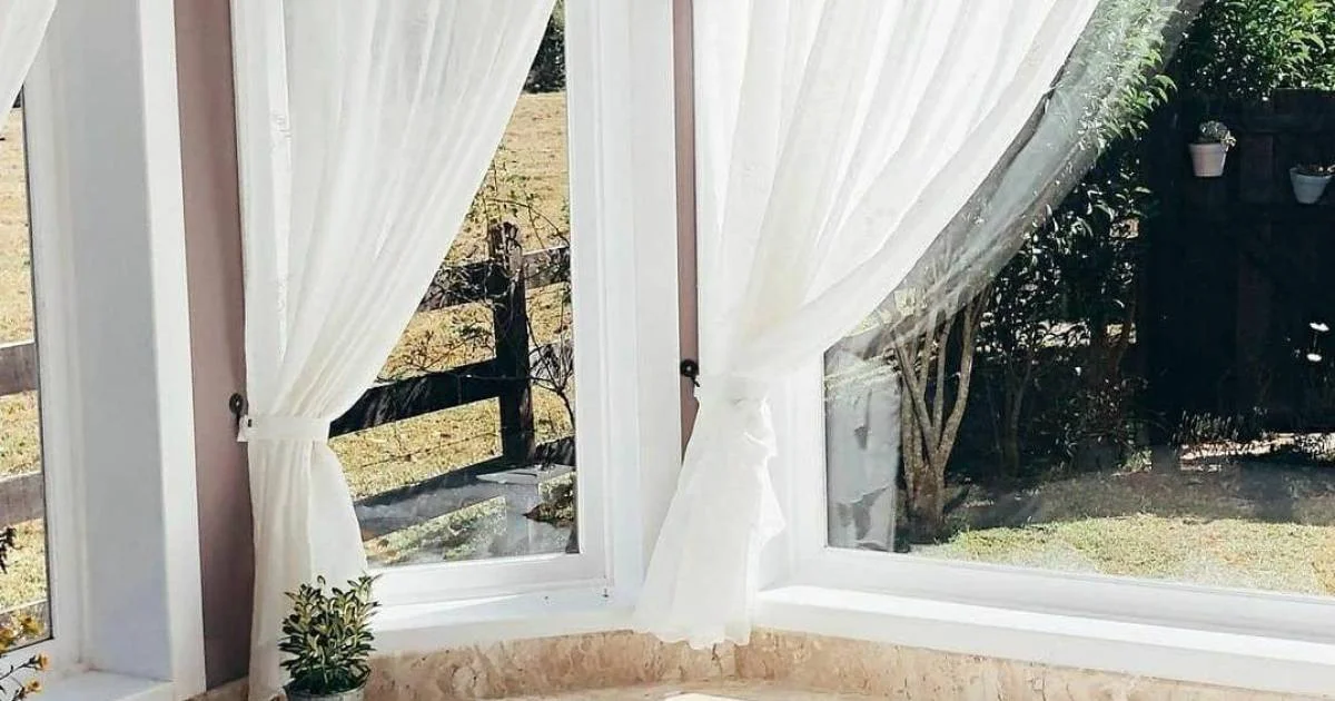 Cortinas brancas e luz natural em suíte da pousada.