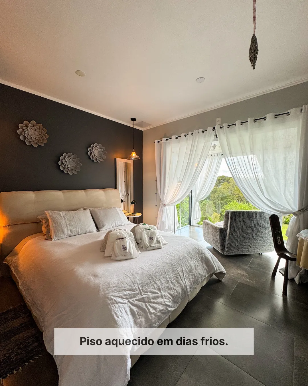 Suíte da Cantos e Encantos com cama de casal, piso aquecido, poltronas junto à janela e vista para o jardim de altitude em Urubici.